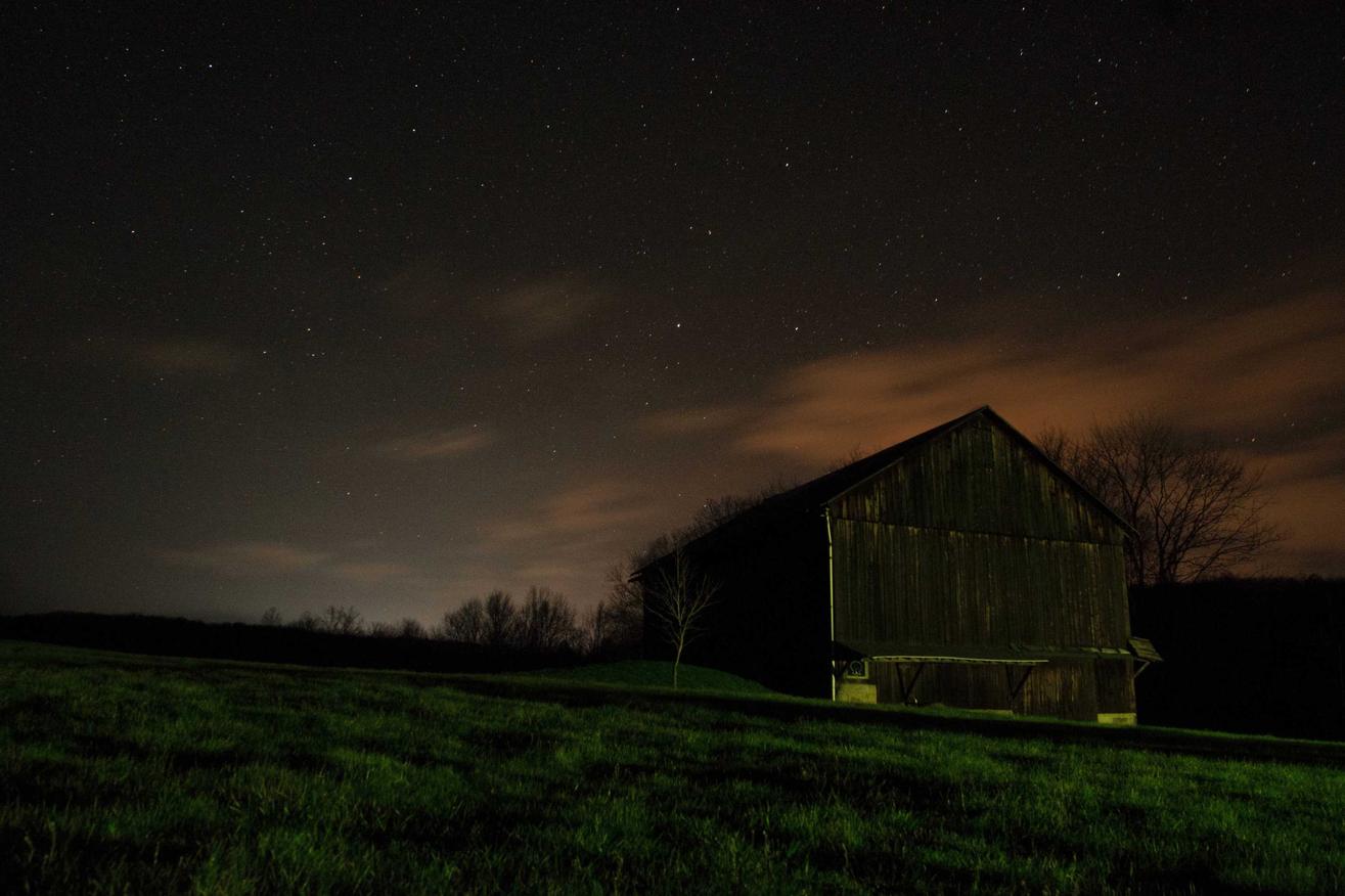 An old barn stands alone in a field at night. The sky is dark with stars shining through.