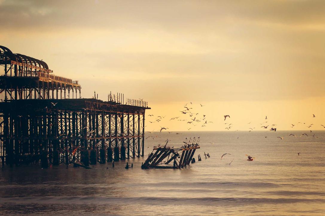 An old pier standing in the ocean with a destroyed section. Birds are flying around.
