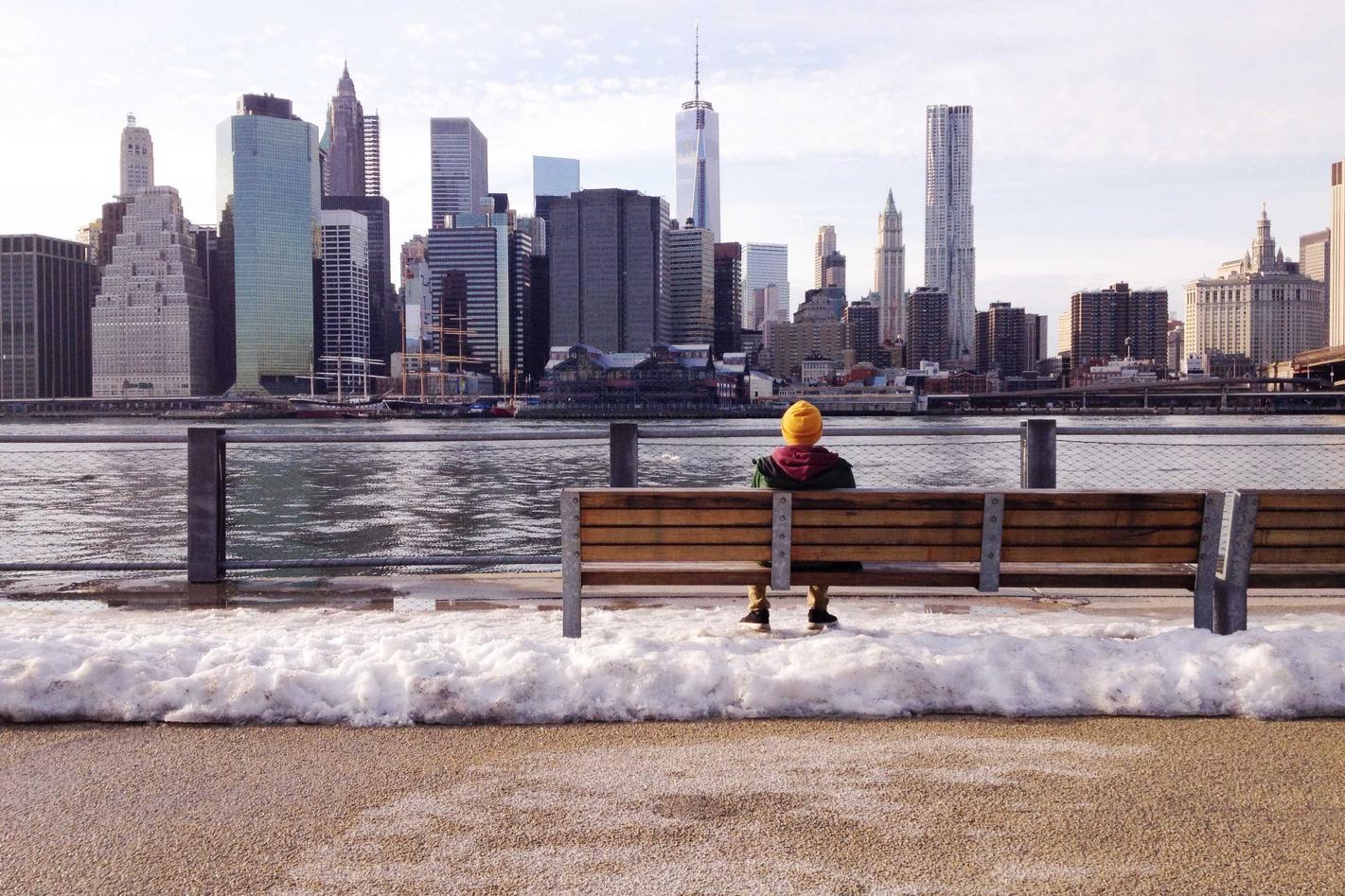 Person sitting on a bench overlooking a city. The city skyline is visible across the water.